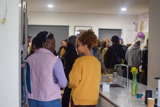 A group of people standing downstairs in a very full YARD. The two people in focus are talking together. One wears a purple top, pink gilet and jeans, with their hair in short twists, pushed back with purple sunglasses. The other wears a bright mustard jumper, with their hair up and glasses.