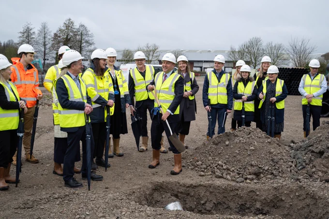 A group of people wearing high-vis jackets and hard hats breaking ground at a building site.  In the front, several are holding spades and a large hole has been dug.
