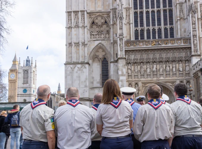 A group of Scouts, in uniform, standing outside Westminster Abbey and looking up towards the west towers.