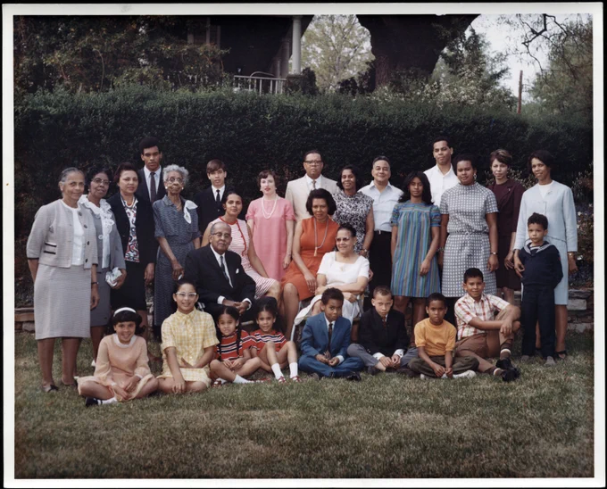 Colour photograph of a family portrait with 28 people posing outdoors on the grass in front of a hedge. 