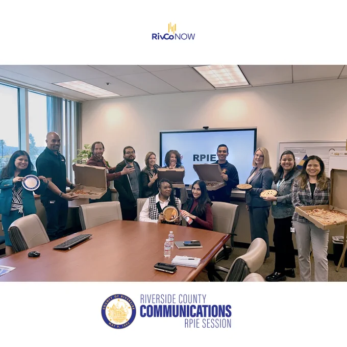 Riverside County Public Information Officers posing together in front of a white screen with the words RPIE in black. Some of the employees are holding pizza pies while others are holding pies.