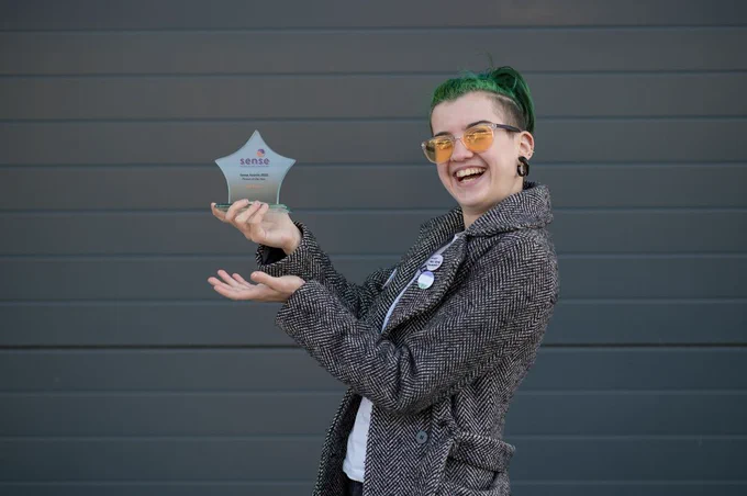 Max rocks bright green hair, orange glasses, a tweed blazer, and chunky black earrings. They smile as they proudly show off a star shaped award from the organization “Sense”.