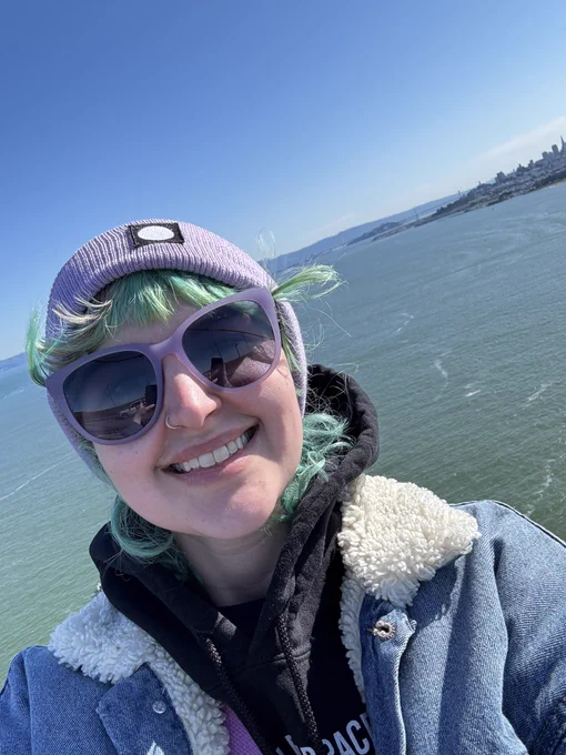 Selfie of Rachael smiling with the bay in the background. They’re wearing a lavender beanie with the Wix Studio logo, lavender sunglasses, a black hoodie, and sherpa lined jean jacket.