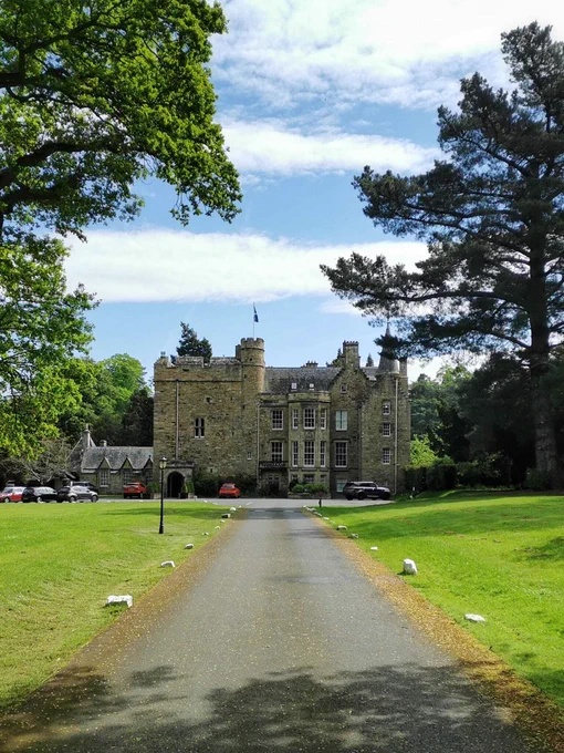 Carberry Tower sits at the end of a long driveway lined with lush green grass and tall green trees.