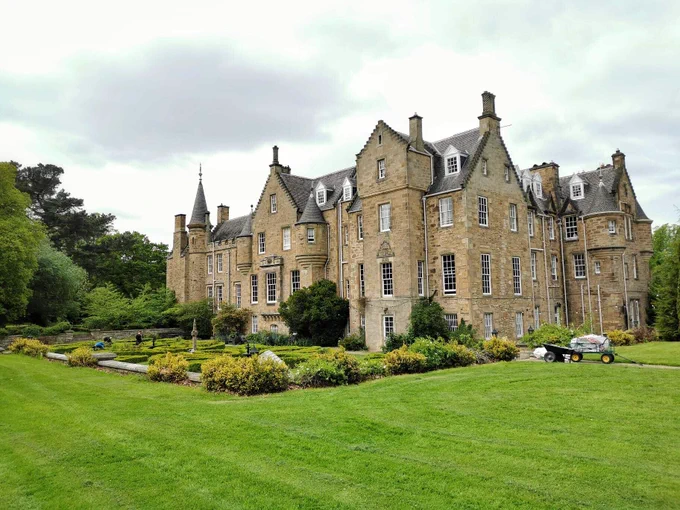 A large country house with lushes green gardens surrounding the exterior. In a few places small turret-like towers can be seen echoing the history of the building.