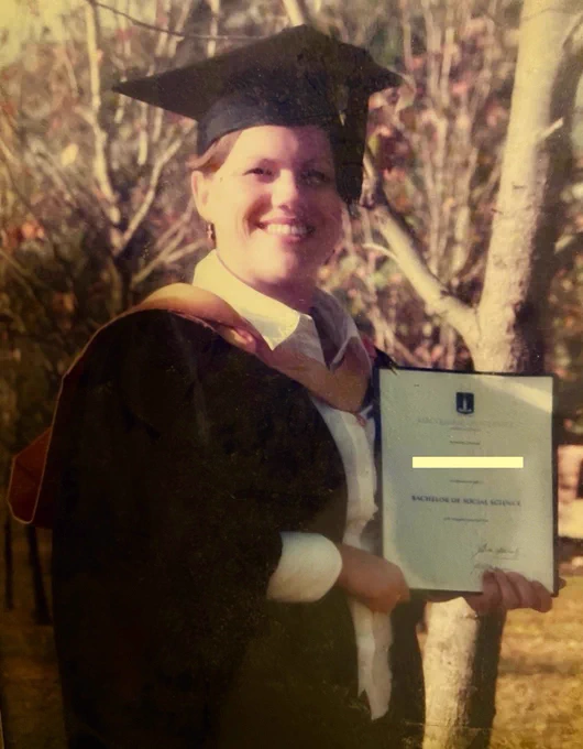 Old discoloured pic of me in a field of trees. I’m proudly wearing a graduation gown and holding my bachelor piece of paper.