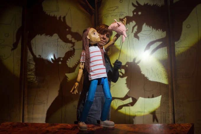 A wooden puppet of a little girl with a female puppeteer handling her. Shadow puppets of wolves in the background.