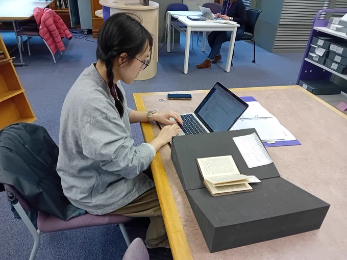 Technology and Medicine student Yunqi He in the map room of the Main Library. She is seated at a desk typing on a laptop, with a book resting beside her on a foam support.