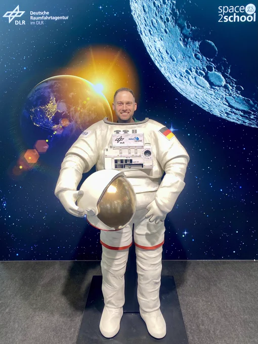 Vor einem großen Bild (wieder drei mal drei Meter) an der Seite unseres Standes, das eine hinter der Erde aufgehende Sonne sowie den Mond zeigt, steht die große Figur eines EVA-Raumanzuges. Man kann sich so dahinterstellen, dass es so aussieht, als würde der eigene Körper im Raumanzug stecken, da nur der eigene Kopf oben aus dem Anzug rausguckt. Auf diesem Bild hier gucke ich grinsend „aus dem All“ in die Kamera.