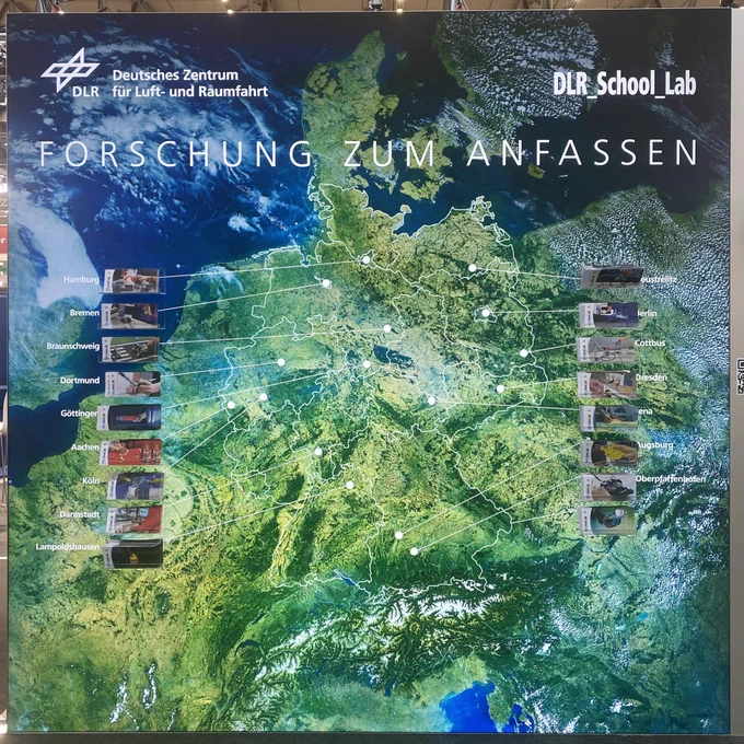 Eine Seite unseres Standes mit einem ungefähr 3 Meter mal 3 Meter großen Satellitenbild Deutschlands. Die Umrisse der Bundesländer sind mit weißen Linken markiert und an allen 16 Standorten der DLR_School_Labs sind weiße Punkte, die durch Linien mit den jeweiligen Flyer-Boxen am Rand verbunden sind. Ungefähr auf Höhe von Dänemark steht unser Slogan „Forschung zum Anfassen“.