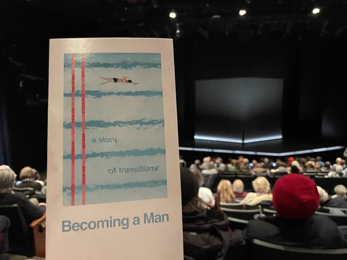 A playbill with the words "A story of transitions - Becoming a Man" is in the forefront of a theatre full of people.