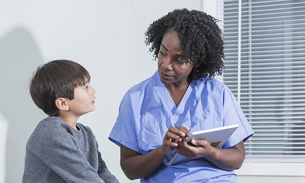 Nurse holding a tablet talks to a young boy. They are both looking at each other as if in conversation.