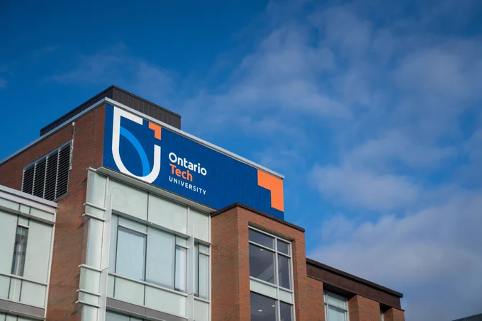 Branding at the top of the Business and Information Technology building shows the university logo on a blue background. The top of the building is shown against a blue sky dotted with fluffy white clouds