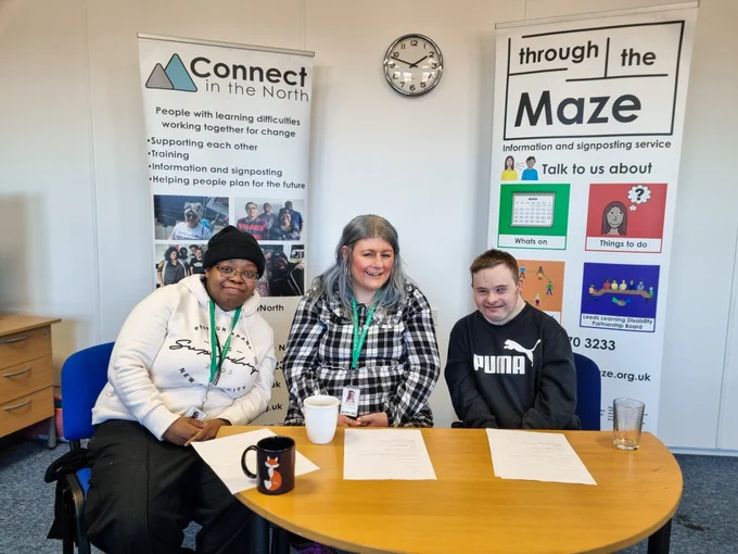 Tania, Vicky and Chris are sat smiling in front of the Connect in the North and Through the Maze banners.