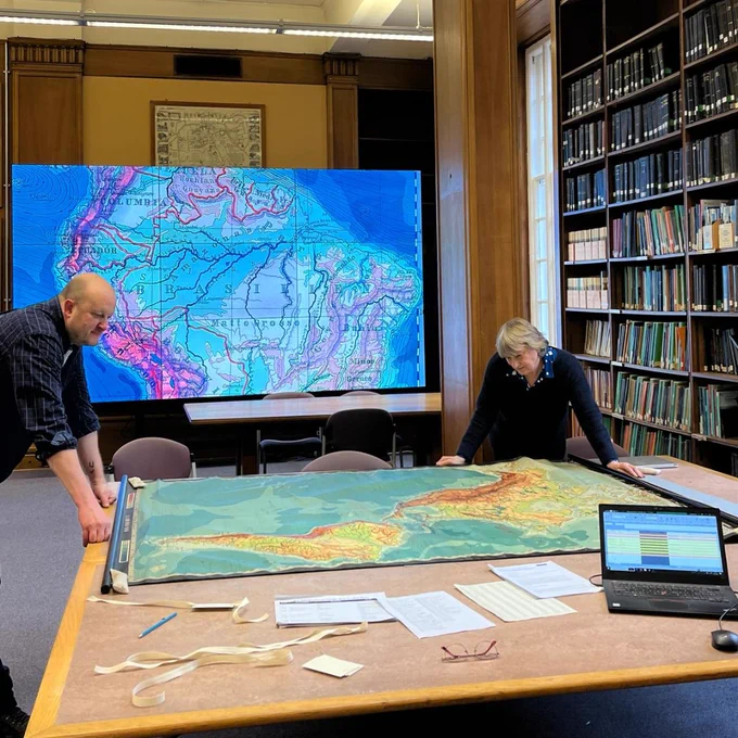 Lise Hopwood and Peter Moody in the map room of the Main Library. In the foreground they are looking at a map on a large table. The map is shown on a large screen in the background.