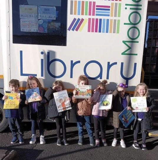 Children holding up their library books outside the mobile library, while sharing their eyes from the sun.