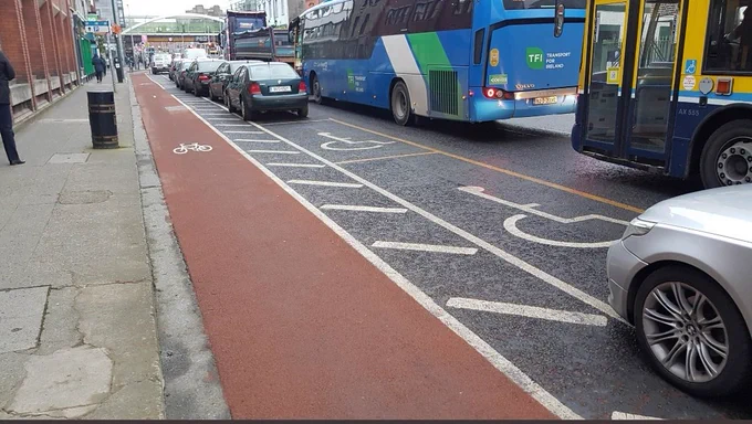 Lombard St Dublin at the back of Trinity college showing 2 vacant disability parking bays wedged in between a busy road with a coach and bus on drivers side no door room for driver. On passenger side is a cycle lane with a narrow buffer zone to protect cyclists from dooring. After the cycle lane is the path with no dropped kerb access. 