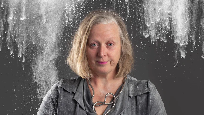 A woman smoking against a grey background wearing a grey top and necklace. There is baking flour falling around her 