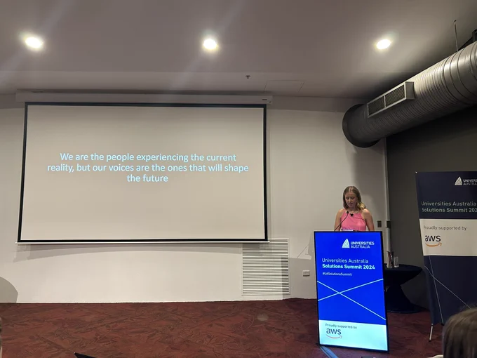 A young woman presenting at the conference, behind her a slide reading “We are the people experiencing the current reality, but our voices are the ones that will shape the future”