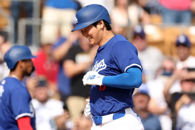 Shohei Ohtani smiles wide as he crosses home following his first Spring Training homer with the Dodgers. He wears a blue Dodgers uniform with white lettering and New Balance batting gloves.