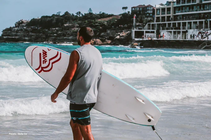 Man with surfboard standing on Bondi beach looking out to sea