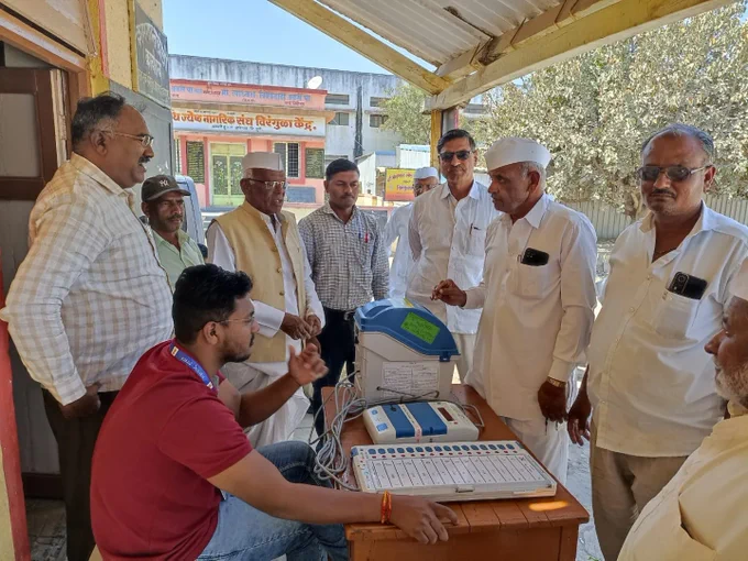 वरील फोटो मध्ये मतदान केंद्रस्तरीय अधिकारी EVM मशीन घेऊन उभे आहेत त्यांच्या भोवती लोकांची गर्दी जमली आहे त्यांना ते EVM मशीनची माहिती सांगत आहे सोबतच प्रात्याक्षिक सुरू आहे.