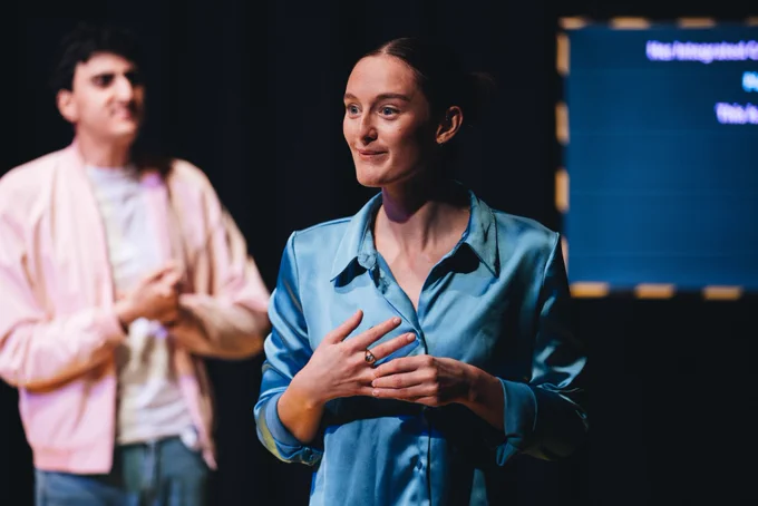 Two actors on stage. One is wearing a pink jacket and white t-shirt, and the other a blue shirt. They are both smiling and, in the background, is a large display screen.