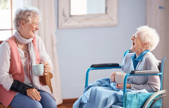 Two women sitting having a cup of tea and laughing. One women is sitting in a wheelchair holding a blue blanket over her lap. 