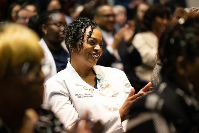 A Black woman smiles amongst a clapping crowd.