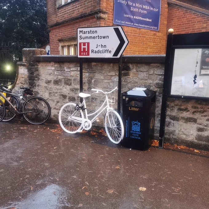 a bicycle spray painted white locked to a road sign on a pavement that reads marston summertown john radcliffe hospital next to a bin and noticeboard with a building in the background