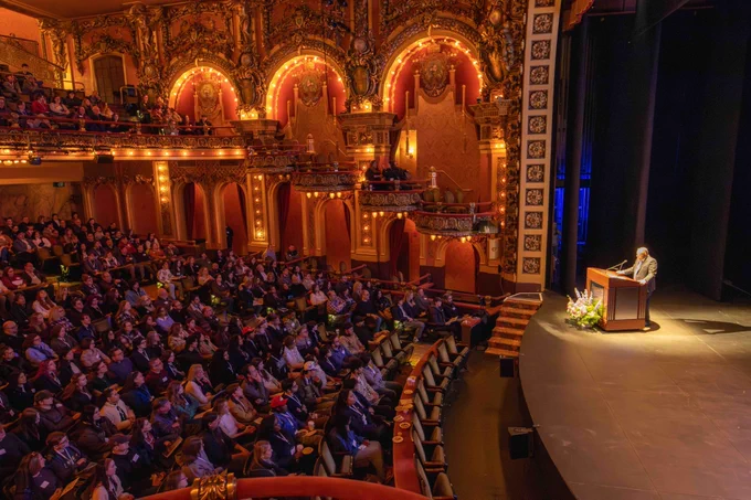 An audience sits in an ornate theatre, listening to a speaker at a podium.