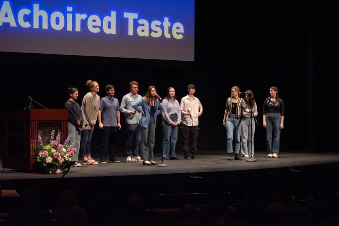A group of performers stand on stage, with one at a microphone.