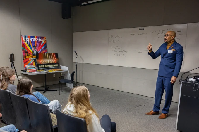 A standing person speaks to people seated in a classroom.