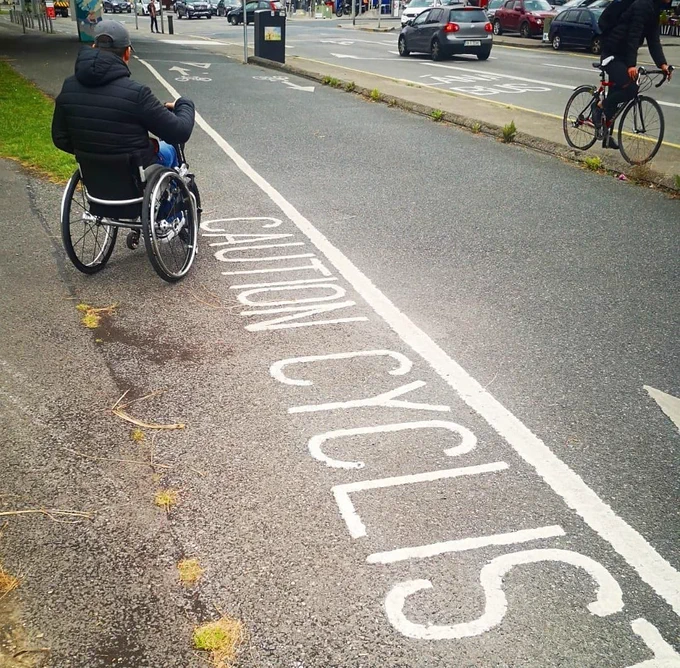 Photo of my husband looking left and right at a contra flow cycle lane along Clontarf which has white painted text saying "Caution Cyclists" showing no cyclists on lane but 1 flying by on the non-designated path for pedestrians. 