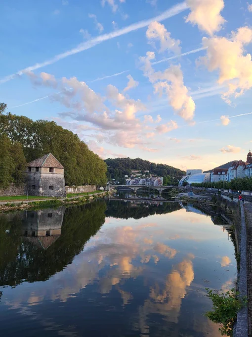 Landscape of Besançon with the river and distant bridge. A few houses are on the river side. The sky is clear and blue with a few clouds. 