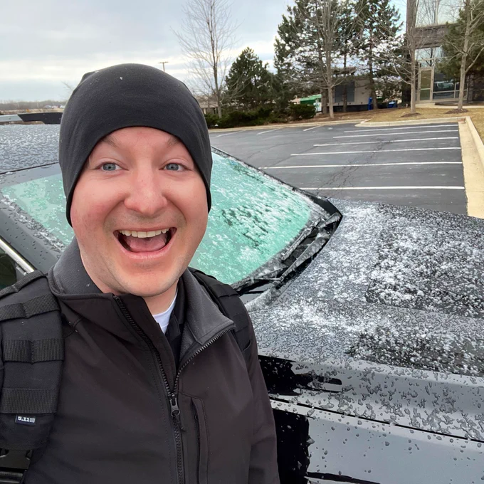 A smiling meteorologist poses with a cheesy smile next to his ice and snow covered vehicle on a cold, windy morning