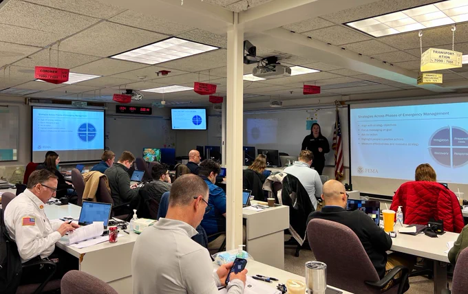 An instructor stands in front of a slide on using social media throughout the disaster lifecycle, at the front of a classroom filled with public information professionals in a social media engagement class