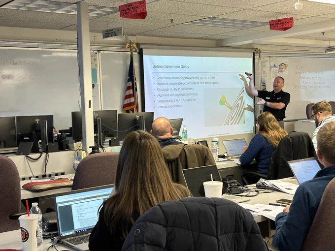 A man in a black polo shirt points to a PowerPoint slide on a screen in front of a classroom of people