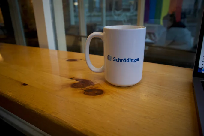 A coffee mug sitting on a wood counter. The mug is branded with the logo for Schrodinger Inc.