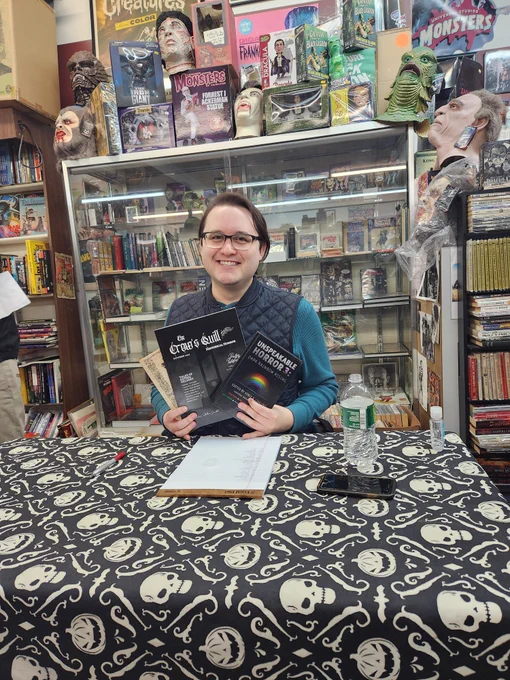 Man with medium length black hair wearing glasses, a teal long sleeved shirt, and a black vest, sitting at a table with a black tablecloth with a white jack o lantern and skull pattern ans holding up a few books