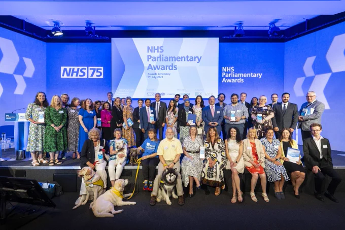 A large group of people are on stage and holding awards. The stage has a screen stating 'NHS Parliamentary Awards NHS75'. They are last year's winners.