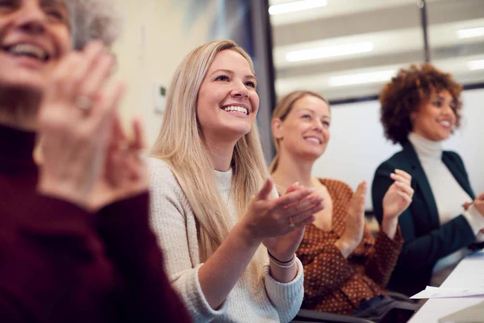 Photograph of four people sitting, smiling and clapping in a conference room. They are all wearing smart casual clothes. The person nearest the camera is slightly out of focus.