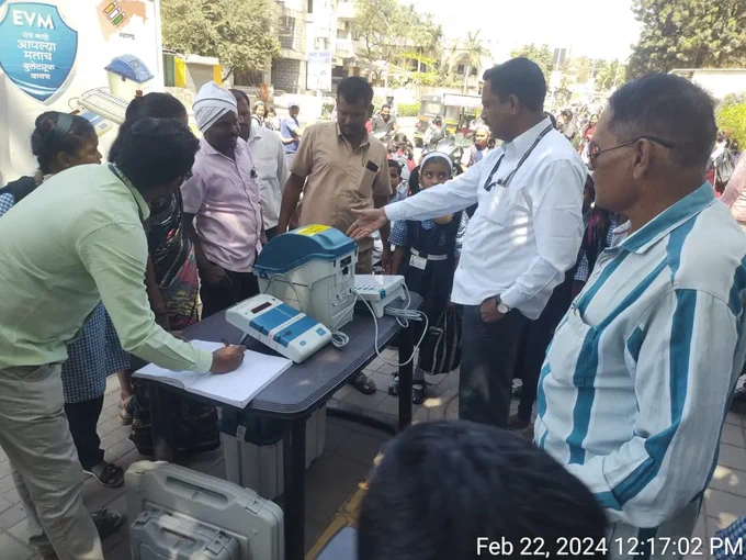 वरील फोटो मध्ये मतदान केंद्रस्तरीय अधिकारी EVM मशीन सोबत उभे आहे आणि त्यांच्या भोवती जमलेल्या गर्दीतील लोकांना ते evm मशीन बद्दल माहिती देत आहे सोबतच प्रात्याक्षिक सुरू आहे.