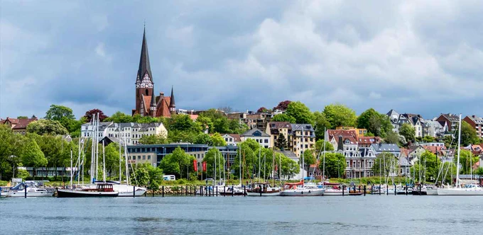 A picture of the skyline of Flensburg showing the sea and buildings between green trees.