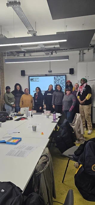 10 adults stand in front of a wall with a screen attached to it. They are all standing close to each other and are smiling towards the camera. There is a table in front of them with laptops cups and pieces of paper dotted around the table