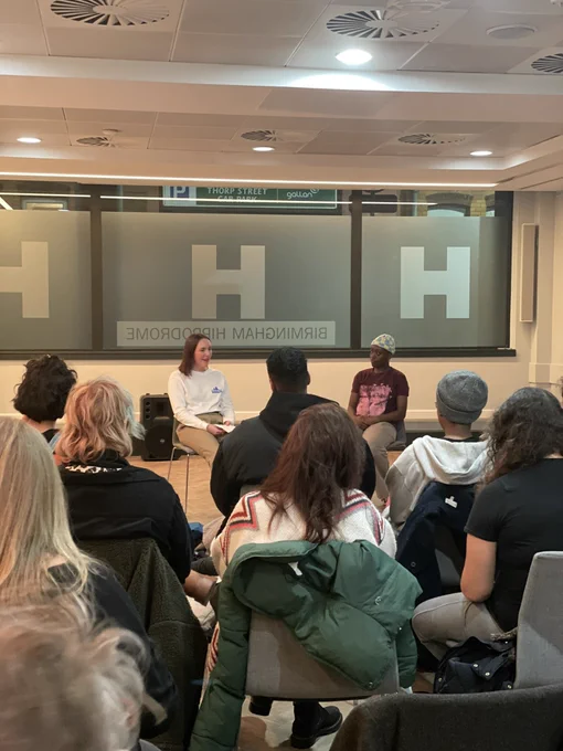 A group of many people sat on chairs looking at two people sharing work