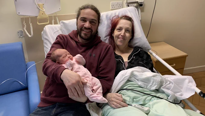 James Poole, in a red hoodie, Emma Loraine, in black, with their baby girl in pink. They are on Emma's hospital bed.