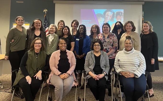 4 women sitting, smiling, 12 women standing, smiling, all are in front of a PowerPoint projection that reads "Collaborative Action for Family Engagement"
