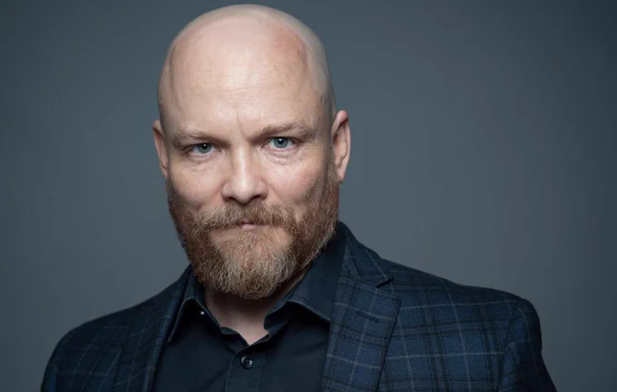 Guy Roberts, a white, bald man with a well-trimmed beard and mustache, wearing a dark blue plaid jacket and a dark shirt underneath it.
