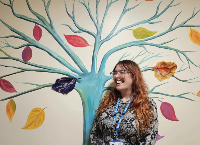 Smiling lady wearing glasses with long red hair and glasses and NHS lanyard standing in front of a brightly coloured tree on a wall in our Eating Disorders Service.